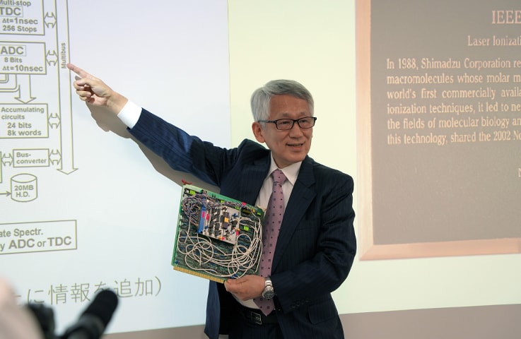 Koichi Tanaka explaining the technology while holding a LAMS-50K circuit board